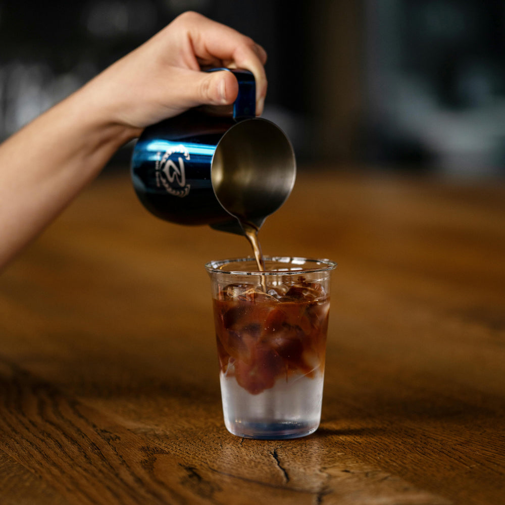 Person pouring a shot of espresso from a blue container into a glass with ice water to make an americano on a wooden surface